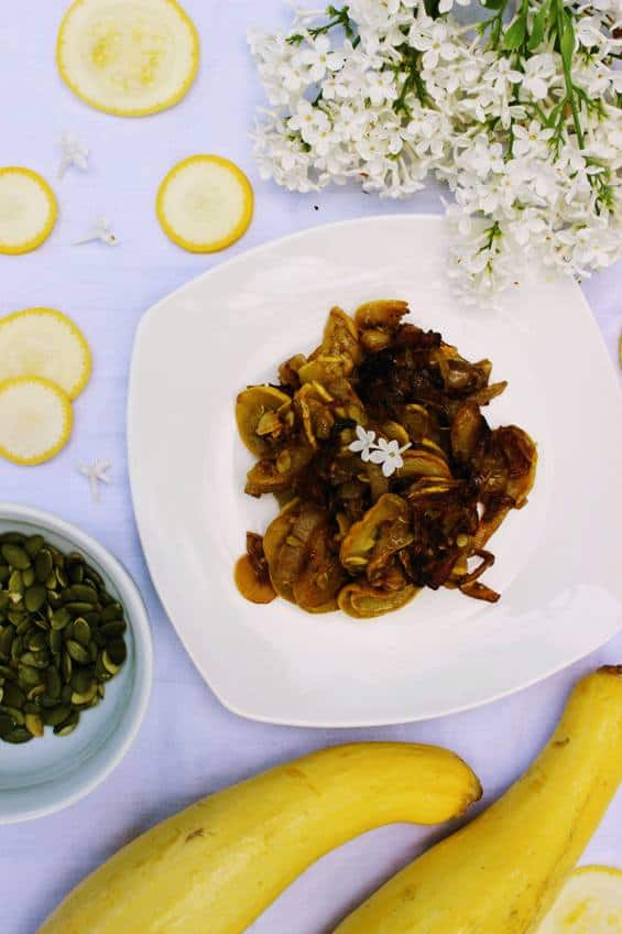 Overhead shot of keto yellow squash and onions on white plate with white tablecloth with 2 whole yellow squash nearby and slices of yellow squash and white lilacs decorating the table