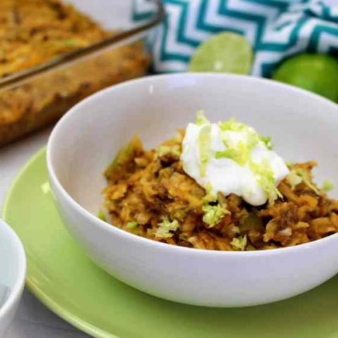 Front view of a serving of keto taco casserole in a bowl, with the pan behind, along with a napkin and limes and a bowl with half an avocado next to it