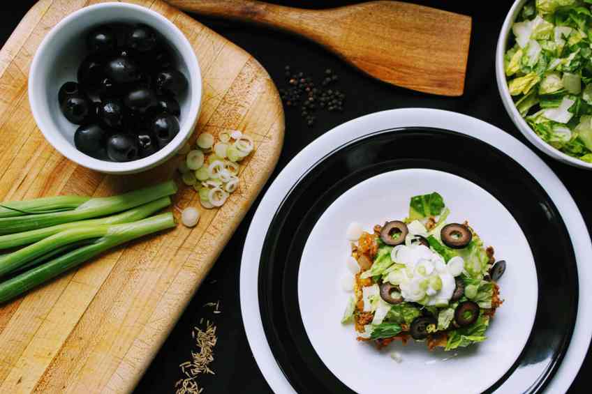 Overhead shot of several things: stacked plates with serving of Keto Mexican Bubble Pizza, cutting board with bowl of olives and partially chopped green onions, and bowl of chopped lettuce